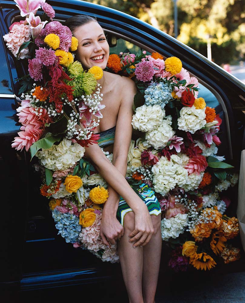 Smiling woman surrounded by vibrant spring flowers stepping out of a car, symbolizing a fresh start and healthy renewal — inspired by HONE&Co’s Spring Clean Hair Kit, featuring the best hairdressers in North Sydney and top products for damaged hair.